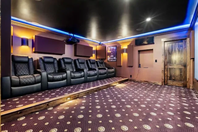 Home theater room in a finished basement with tiered leather recliners, ambient LED lighting, acoustic wall panels, and patterned carpet flooring.