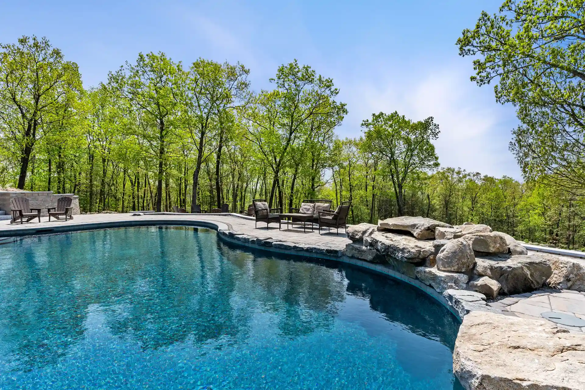 A serene poolside view featuring natural stone accents, a seating area with chairs, and lush green trees surrounding the pool area.