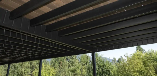 View from below showing the underside of a deck with black metal beams and brown composite decking boards, supported by vertical posts.