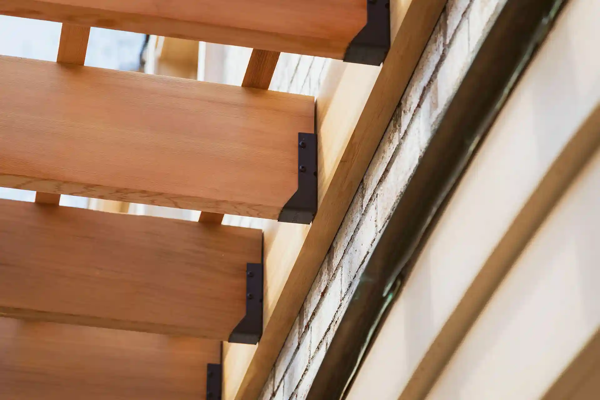 Close-up view of wooden beams with metal brackets attached to a brick wall and siding, showing the underside structure of a deck or pergola. 
