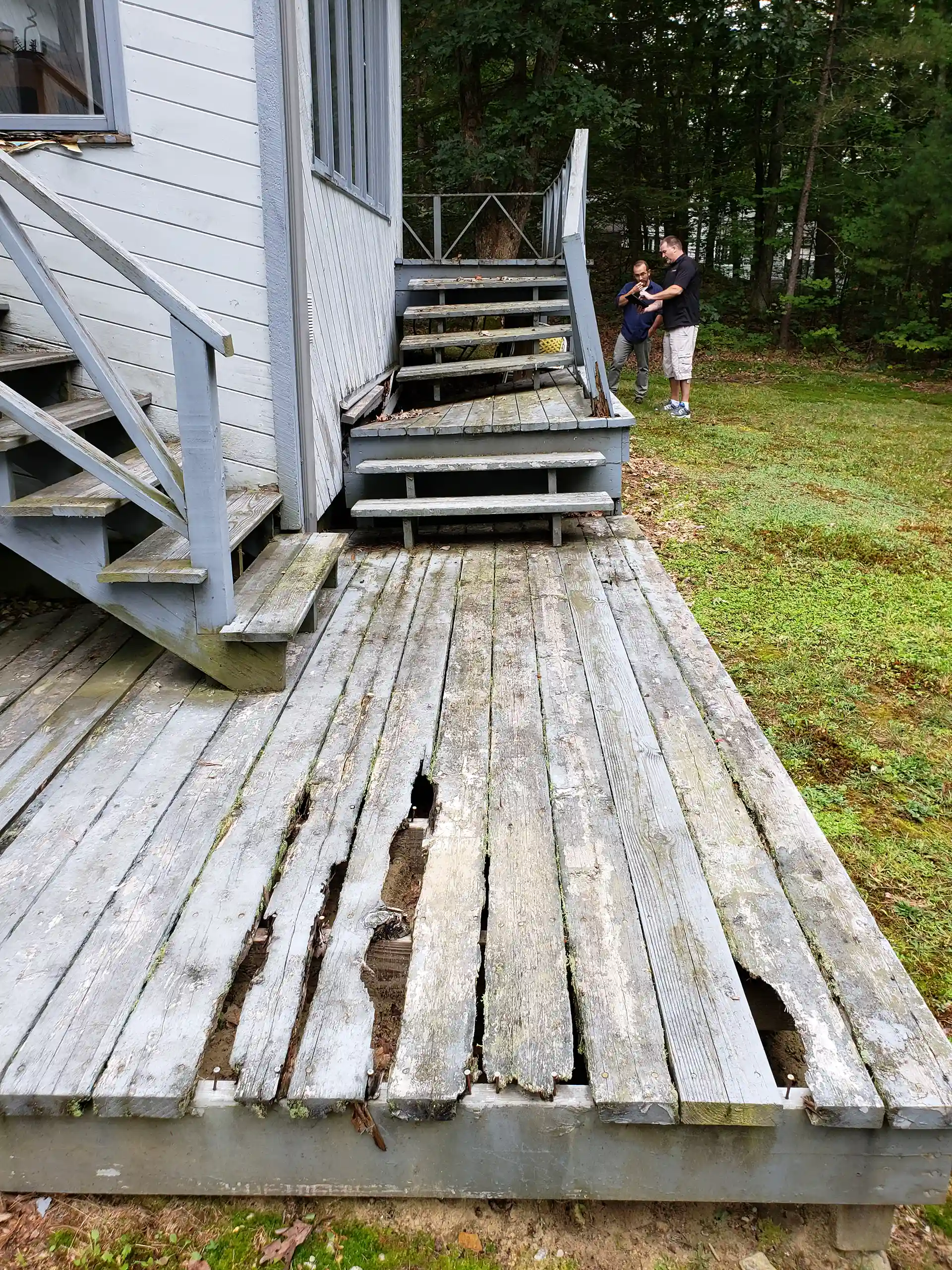 Wood deck with missing and broken boards, stairs leading to the deck, a grass lawn surrounding the area, and a man holding a child standing near the deck in the background. 
