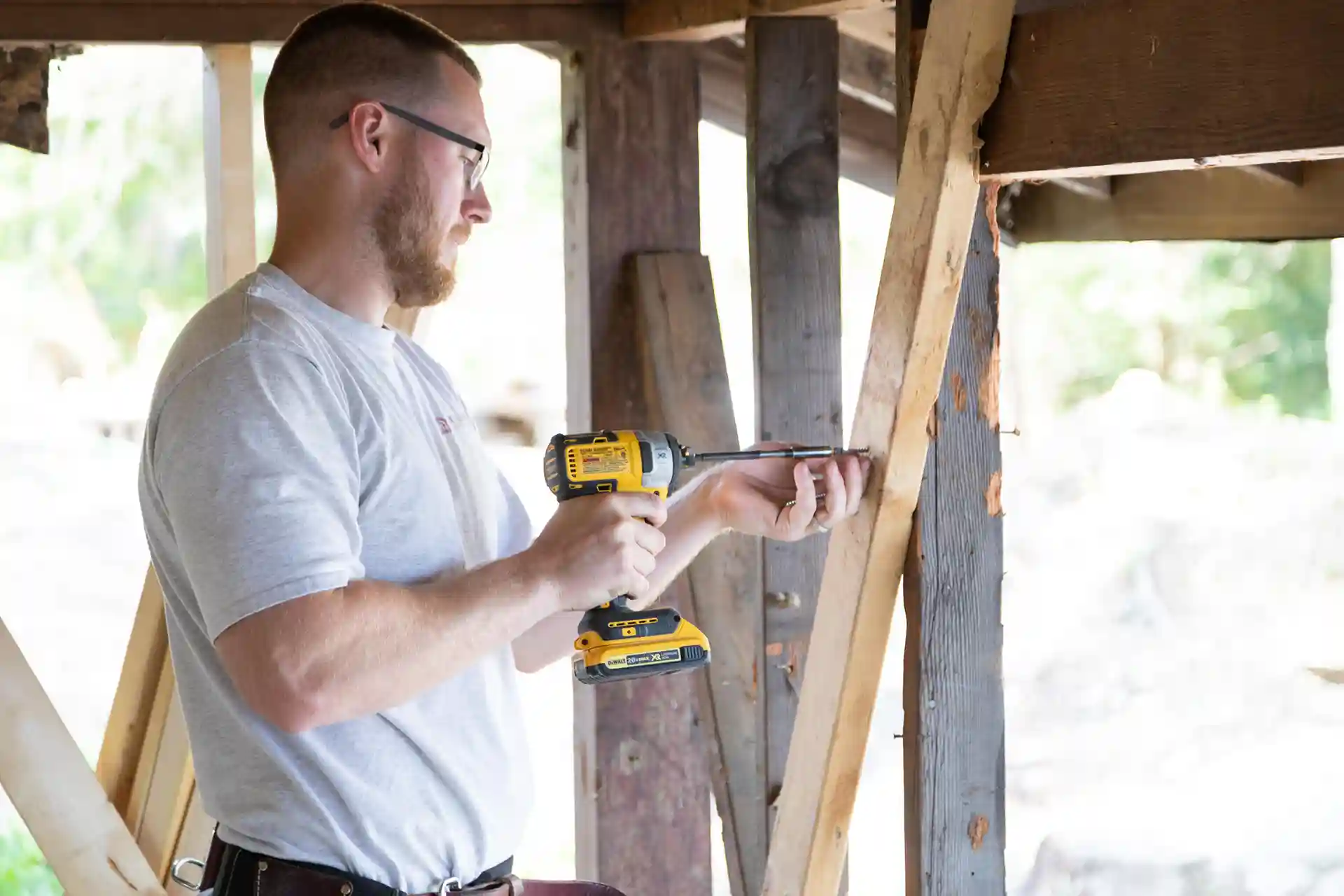 Man using a cordless drill to fasten a screw into a wooden post under a deck during construction or repair work. 