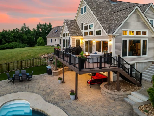 Modern home with elevated deck overlooking a paved patio, outdoor seating, and a pool at sunset.