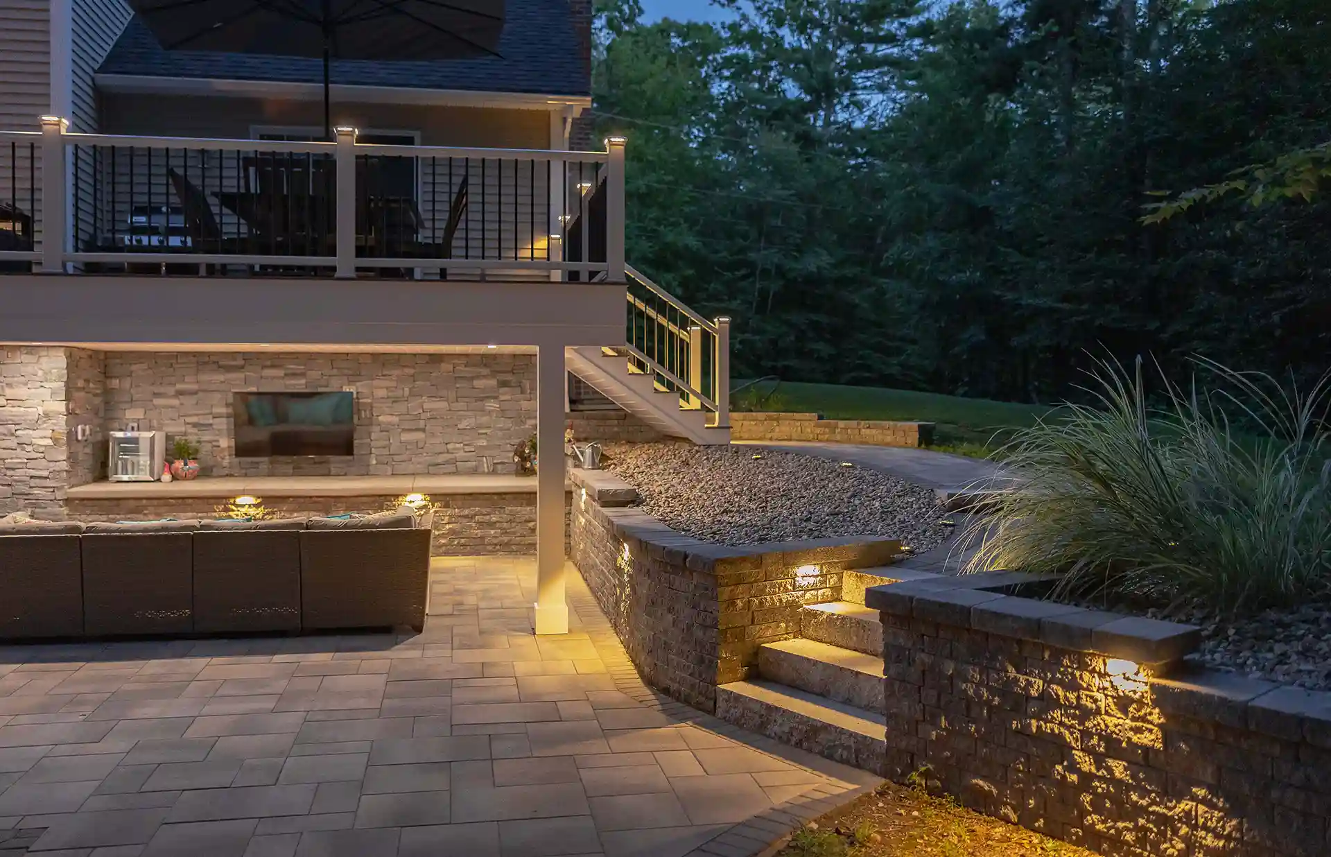 Basement patio at dusk with stone walls, built-in outdoor TV, recessed lighting, seating area, and stairs leading to upper deck.
