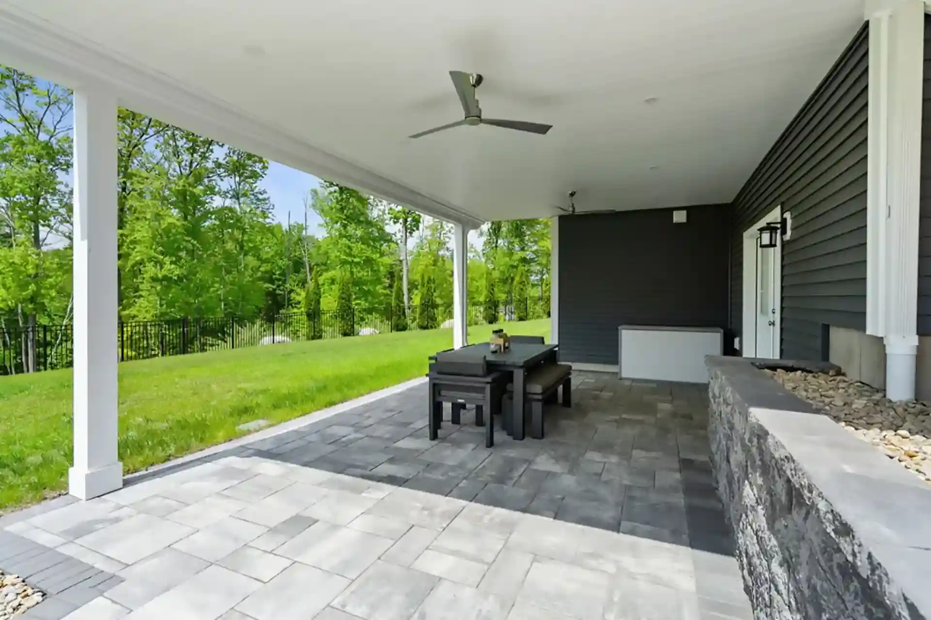 Covered basement patio with ceiling fans, dining table, stone wall planter, and view of green lawn and trees.
