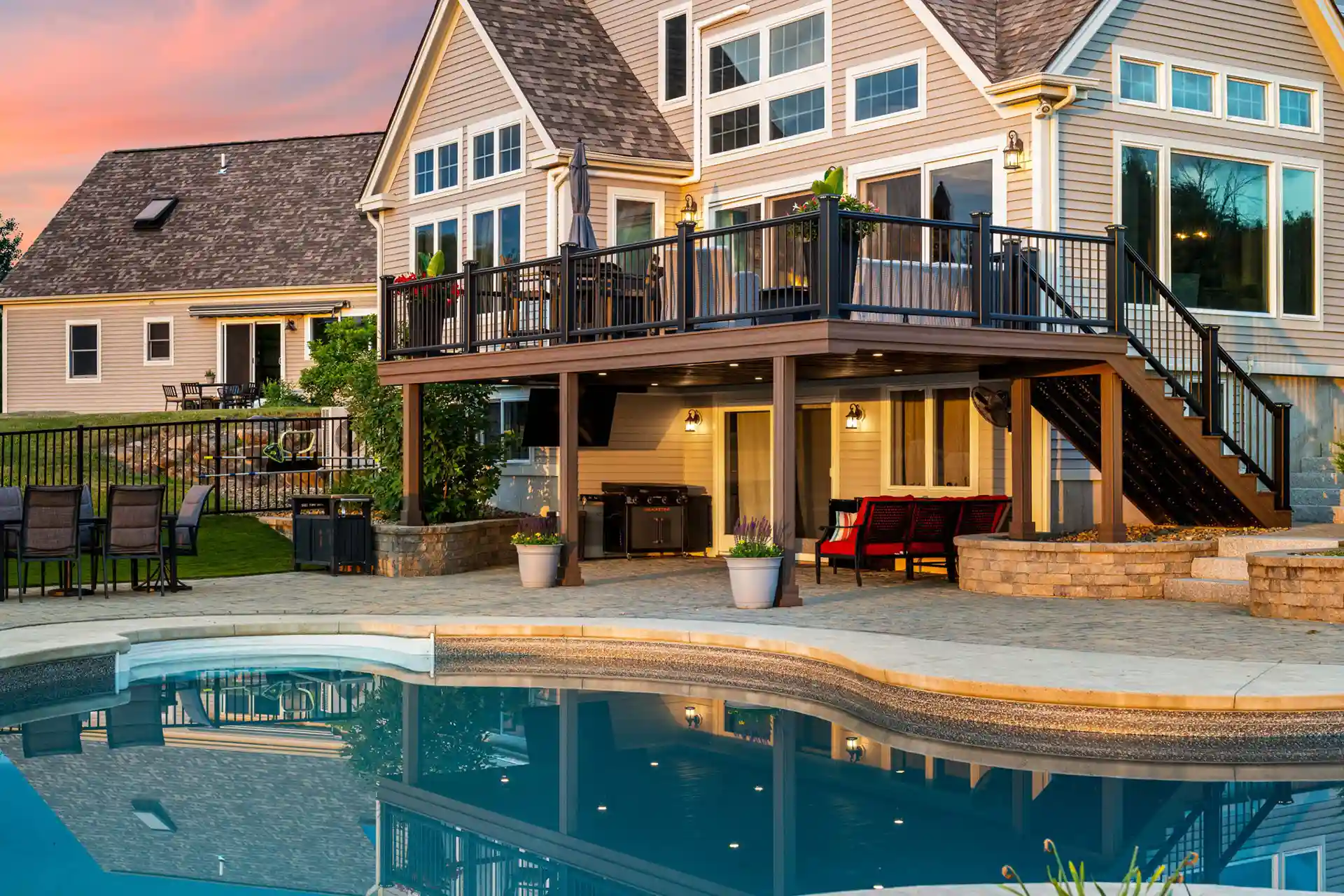 Basement deck with black railing overlooking a paved patio and swimming pool, attached to a modern house at sunset.
